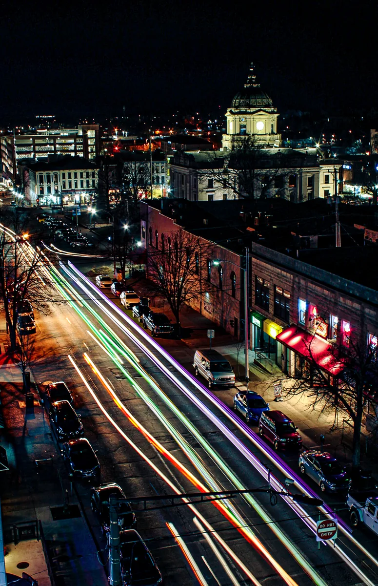 A city street at night from above, headlight trails running past lit shopfronts.