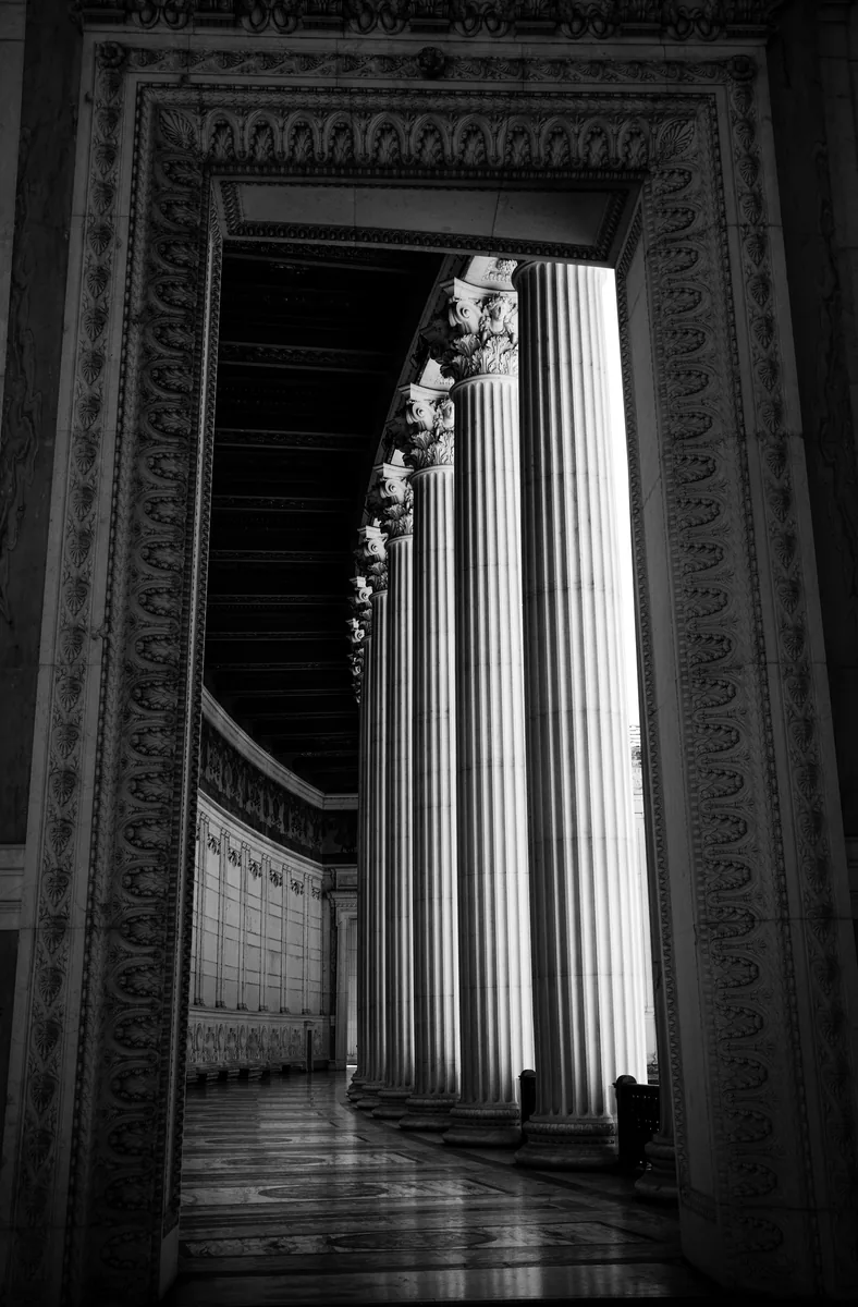 Through an ornate doorway to a sunlit colonnade beyond, in black and white.