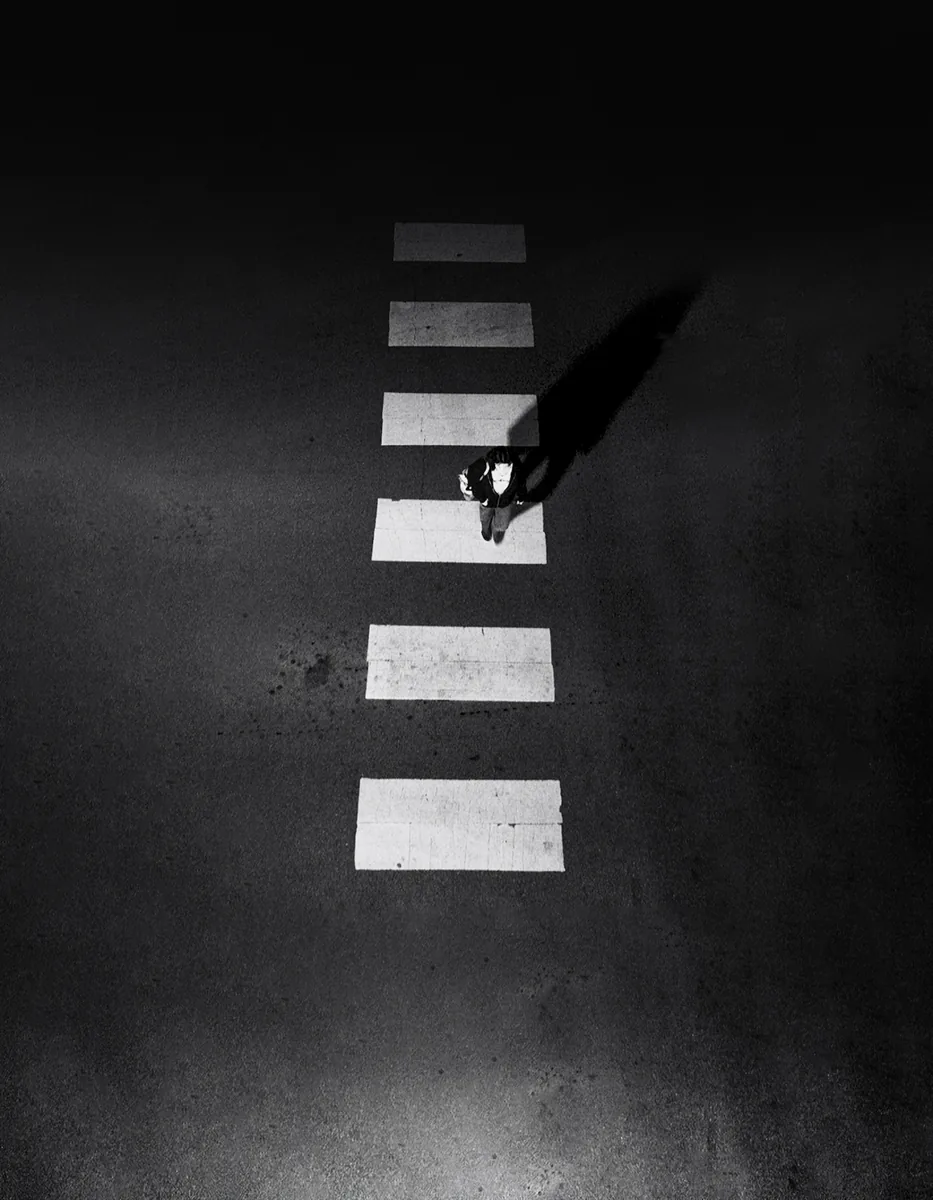 A lone figure crossing a bright zebra crossing on dark asphalt, shot from above.