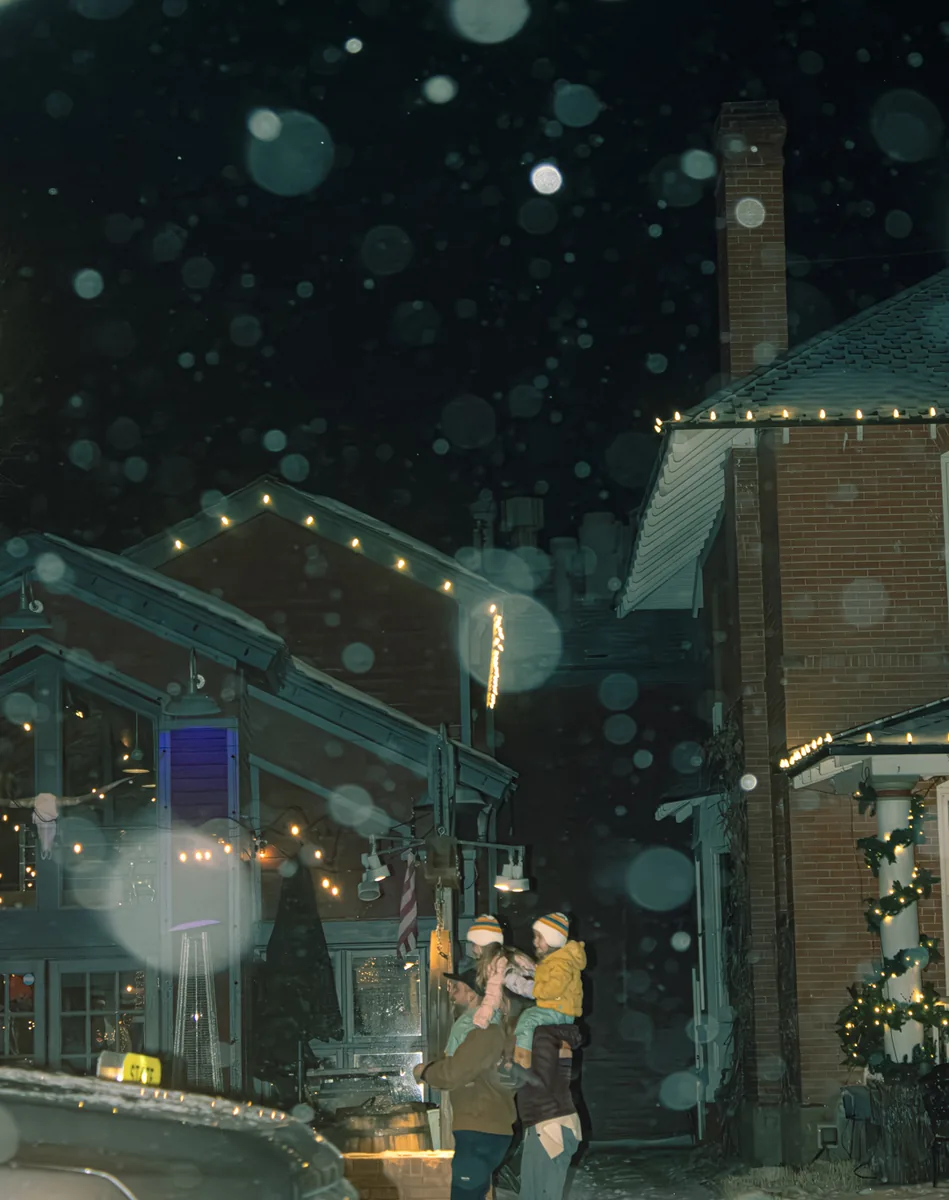Snow falling through string lights over a shop-lined street at night.