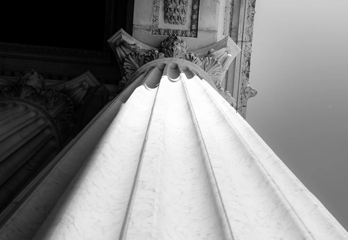 Looking up the fluted shaft of a stone column to its capital, in black and white.