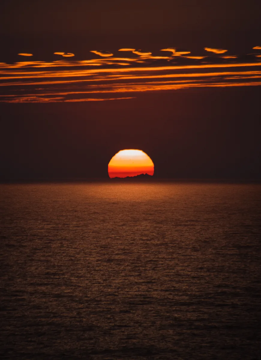 A deep red sun half set on the sea horizon under bands of orange cloud.
