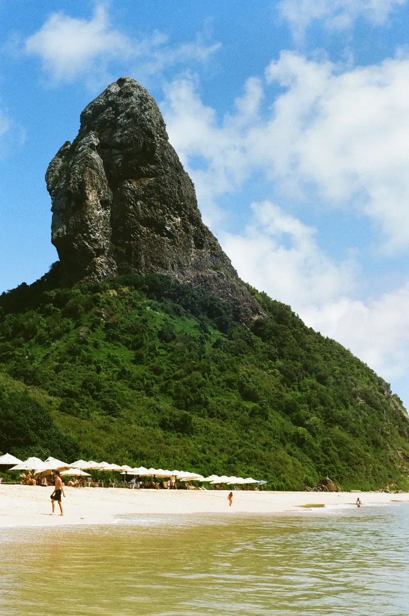 The rock peak above Fernando de Noronha, seen across a beach of white parasols and clear shallows.