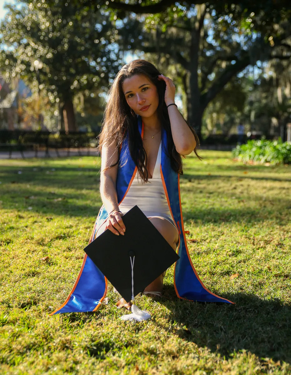 A graduate in a stole kneeling on the grass under live oaks, cap set down in front, University of Florida.