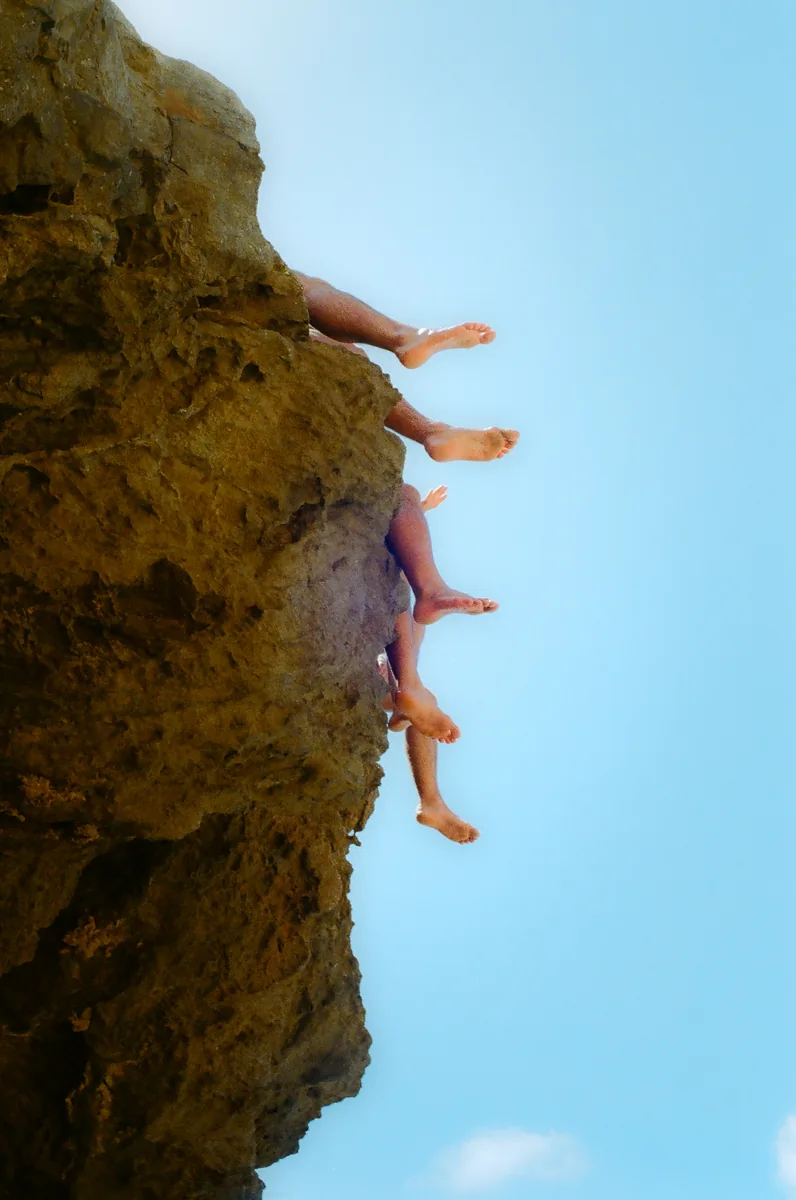 Bare feet and legs dangling over the lip of a sunlit cliff against a blue sky, Fernando de Noronha.