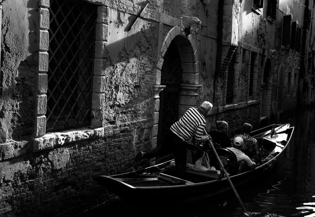 A gondolier poling down a narrow Venetian canal, in black and white.