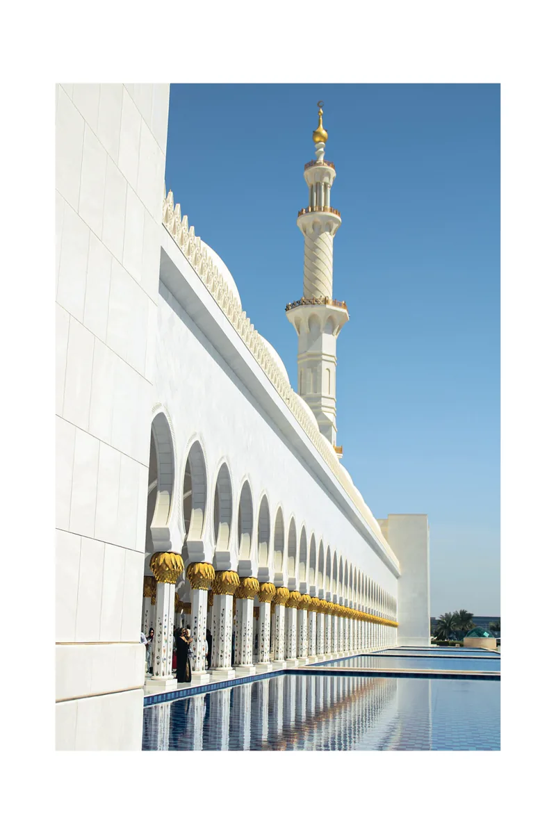 The arcade and minaret of the Sheikh Zayed Grand Mosque running beside a still reflecting pool.